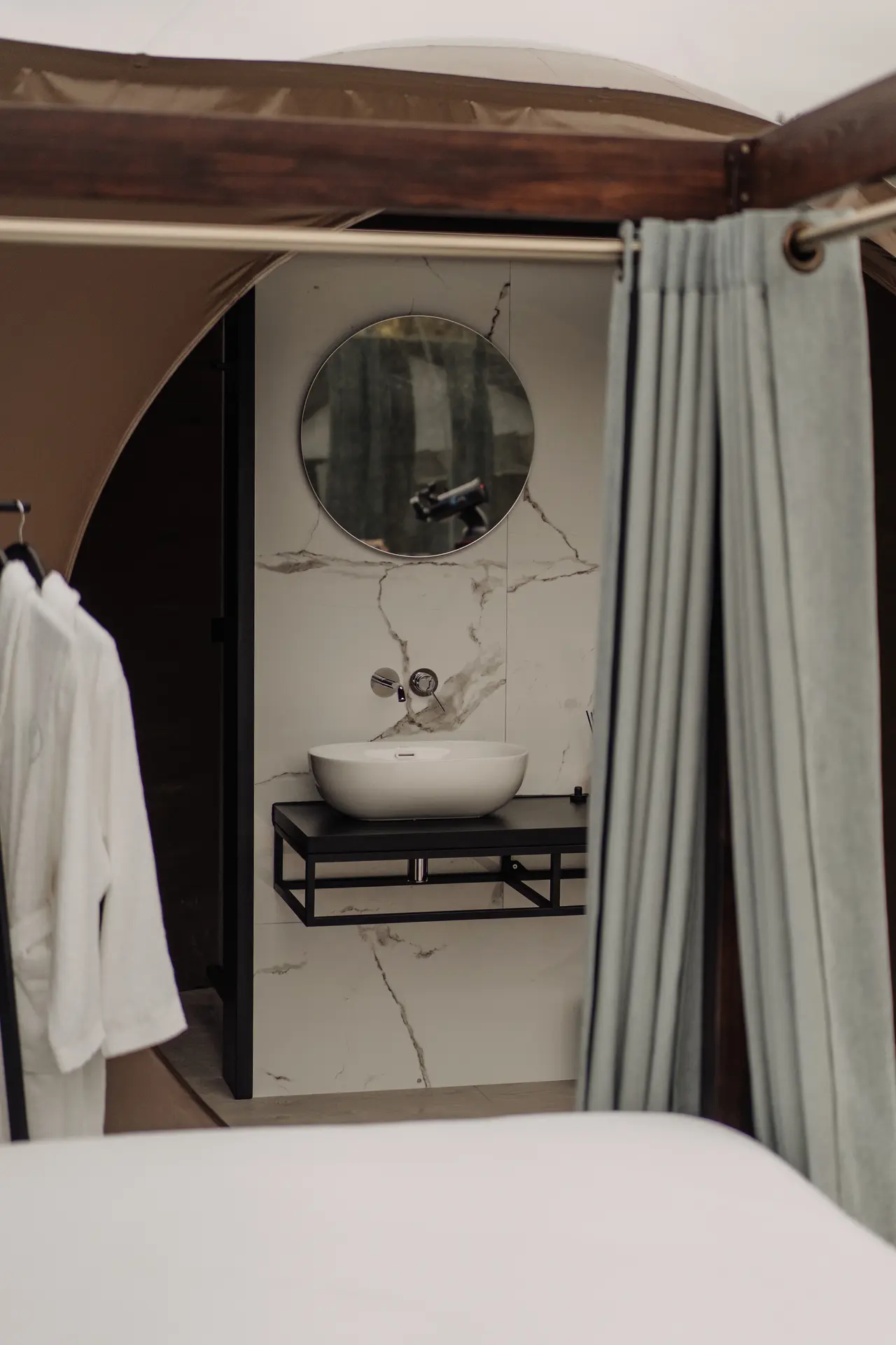 Vista de un baño con paredes de mármol blanco, lavabo blanco sobre un mueble negro, espejo redondo y grifería plateada.