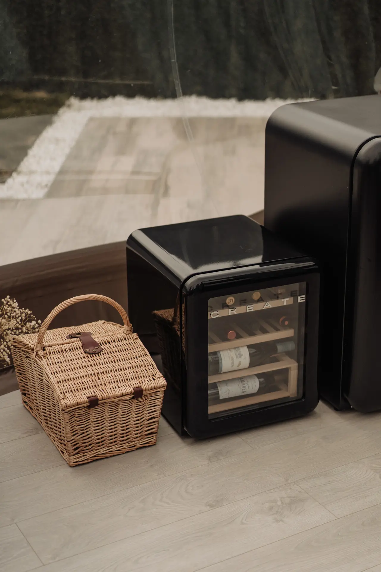 Cesta de mimbre y minibar de vinos negro sobre suelo de madera clara, con vista a un exterior borroso.