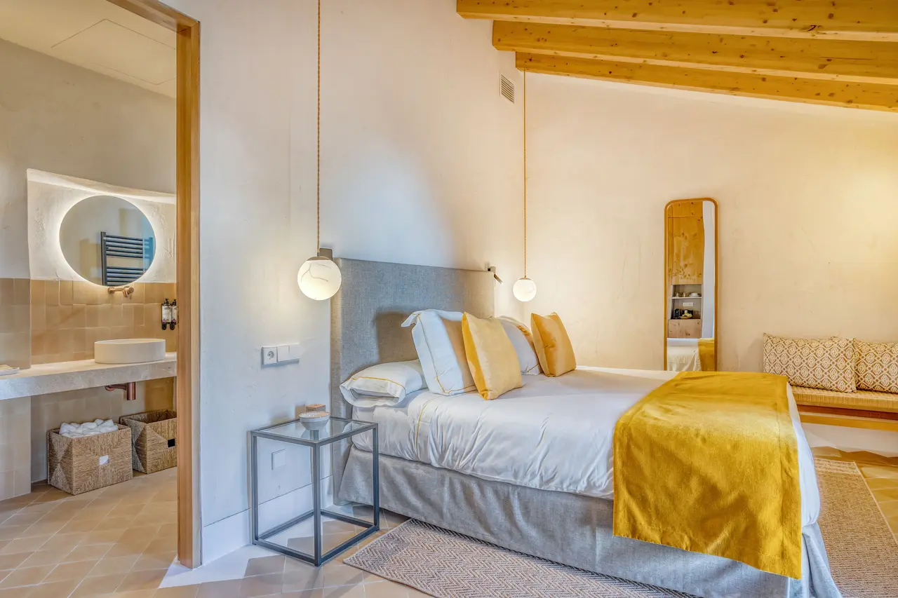 Vista de una habitación con cama grande vestida de blanco y amarillo, mesita auxiliar, sofá al fondo y puerta abierta
