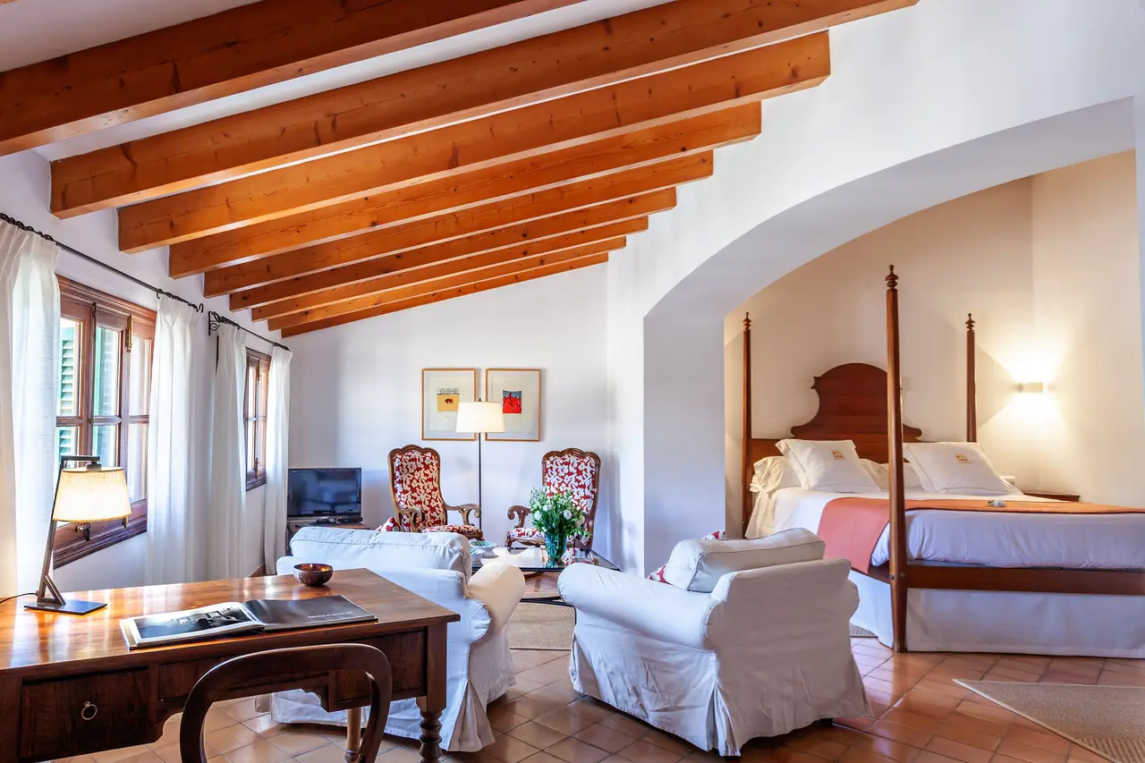 Vista de una junior suite con techo de vigas de madera, cama de dosel, sofá y sillones blancos, escritorio de madera