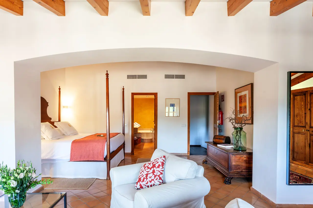 Vista de una junior suite con cama de dosel, sofá blanco, vigas de madera en el techo y una puerta abierta que muestra
