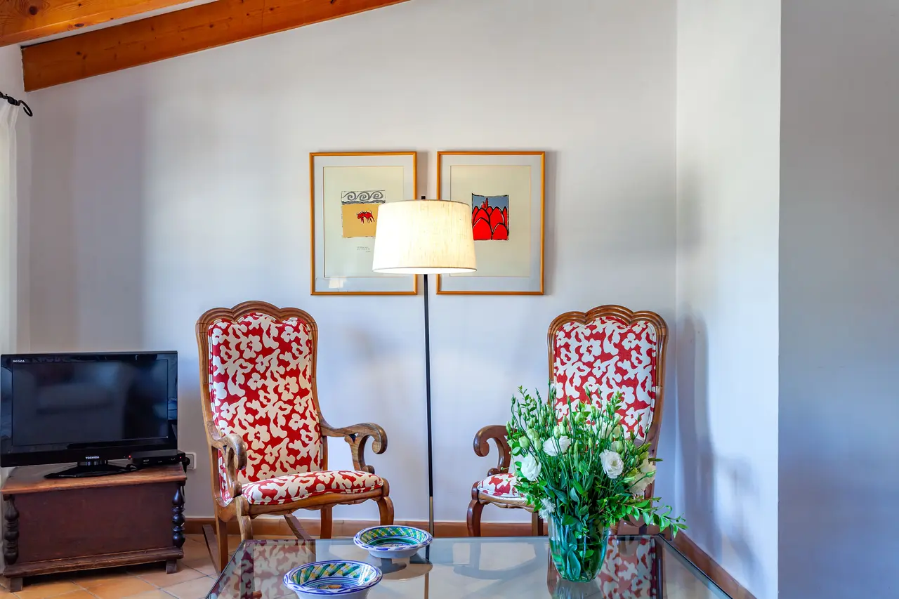 Interior de una habitación con dos sillones tapizados en rojo y blanco, una mesa de cristal con un jarrón de flores