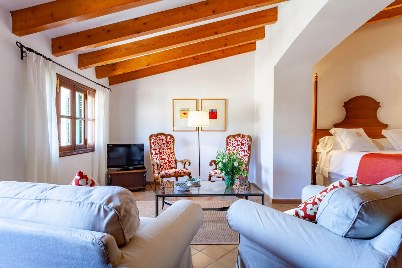 Vista de una junior suite con techos de vigas de madera, dos sofás claros en primer plano, mesa de centro con flores