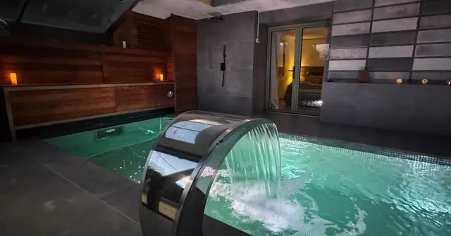 Piscina privada climatizada en interior con agua turquesa, cascada de acero inoxidable en primer plano, paredes de hormi