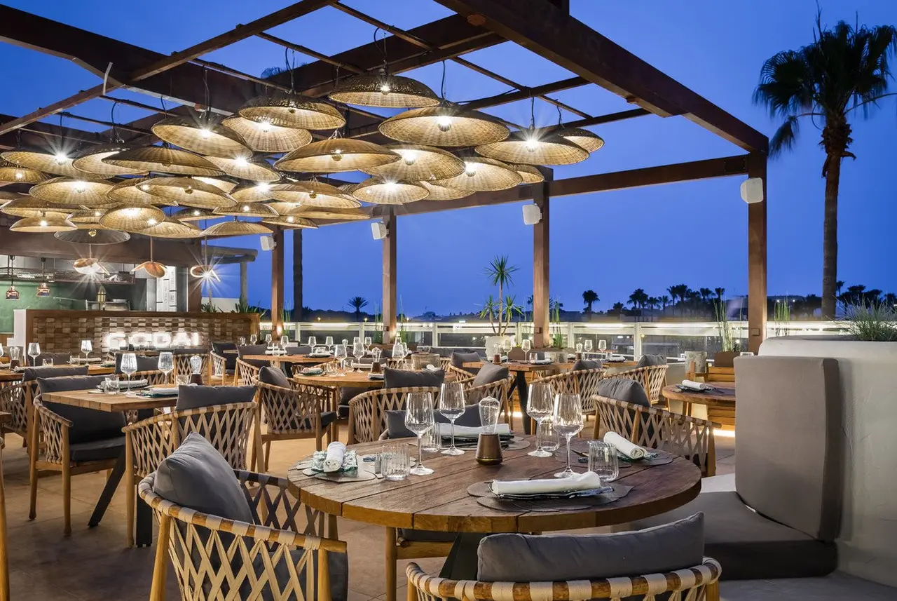 Terraza exterior de restaurante al atardecer con mesas redondas de madera, sillas de mimbre y numerosas lámparas