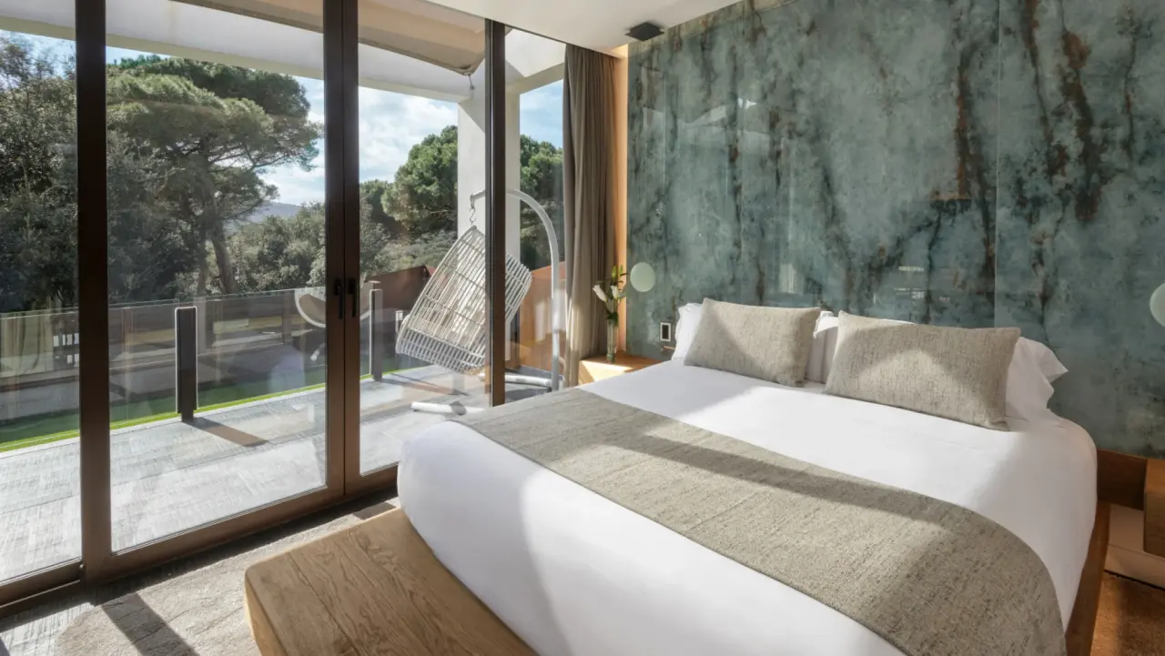Dormitorio luminoso con cama grande, pared de mármol verde y grandes ventanales que dan a una terraza con hamaca