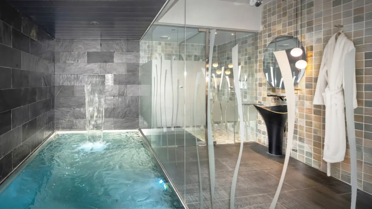 Piscina privada interior con cascada de agua en una suite con ducha, lavabo y albornoz colgado.