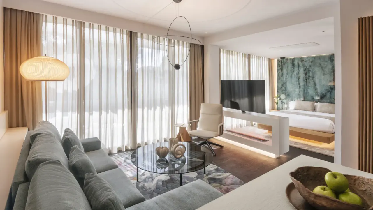 Vista de una suite con sofá gris, mesa de centro, sillón y gran ventanal con cortinas, al fondo se ve
