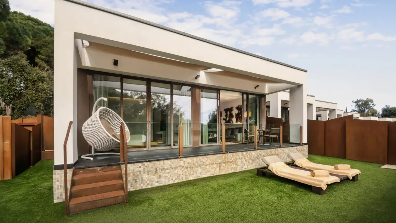 Fachada exterior de una suite moderna con terraza, césped y dos tumbonas en primer plano.