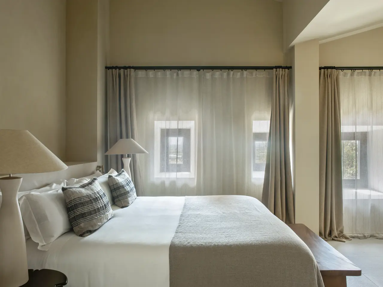 Habitación con cama grande vestida de blanco, cojines decorativos, dos lámparas de mesita de noche, cortinas claras