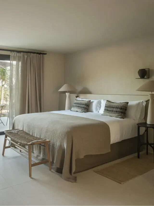 Habitación con cama king size vestida con colcha beige, cojines a rayas, dos mesitas de noche con lámparas, banco