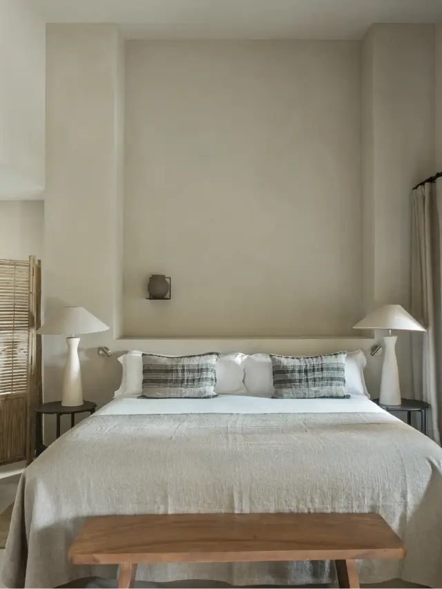Habitación con cama king size vestida en tonos neutros, dos mesitas de noche con lámparas blancas, banco de madera