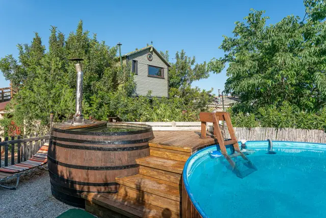 Jacuzzi exterior de madera con escalera, ubicado en una terraza con un barril de agua caliente y una casa de madera