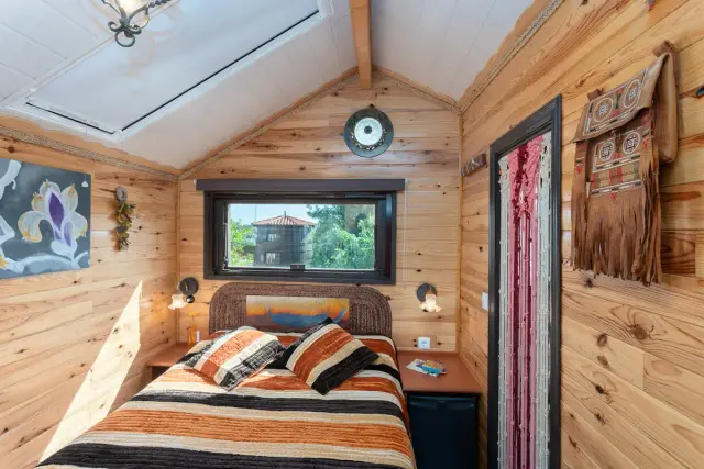 Interior de una cabaña de madera con techo abuhardillado, una cama grande con colcha y almohadas a rayas naranjas