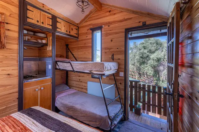 Interior de una cabaña de madera con literas metálicas, una cama en primer plano, cocina compacta a la izquierda