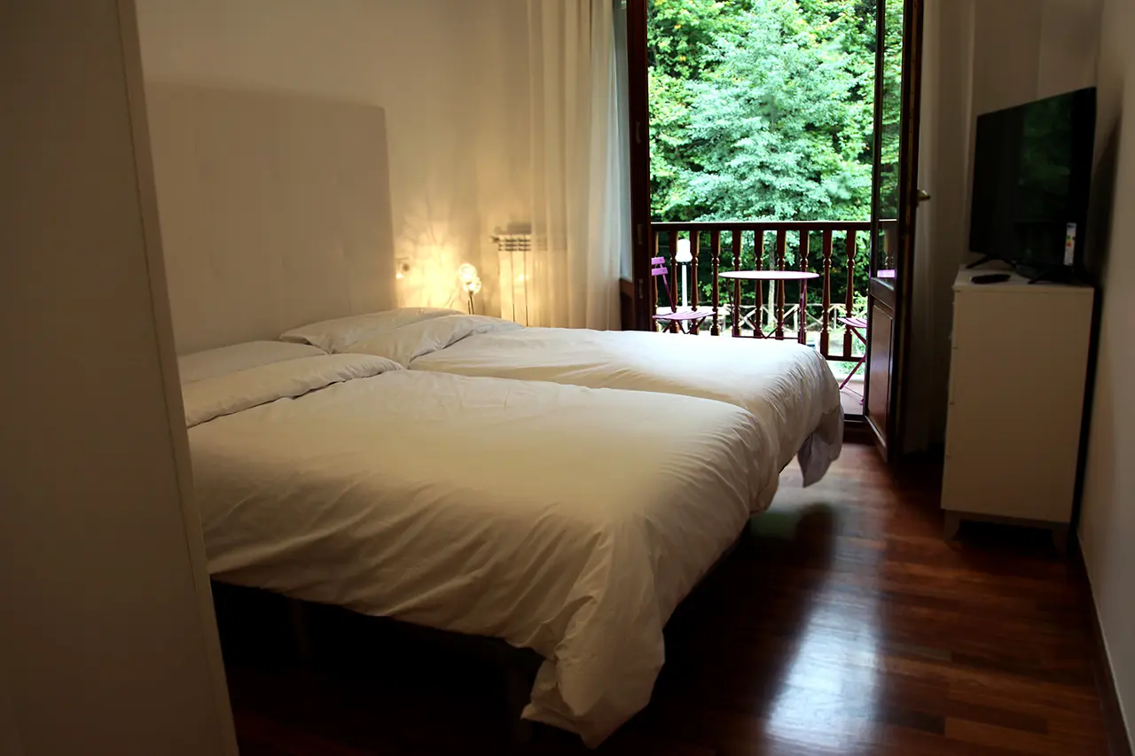 Habitación con dos camas individuales unidas, ropa de cama blanca, cabecero claro, suelo de madera, televisor