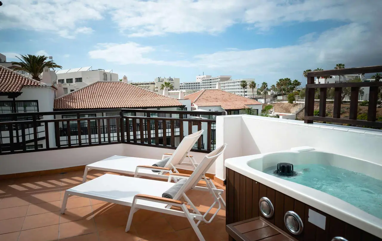 Terraza privada con jacuzzi, dos tumbonas blancas y vistas a edificios y palmeras bajo un cielo nublado.