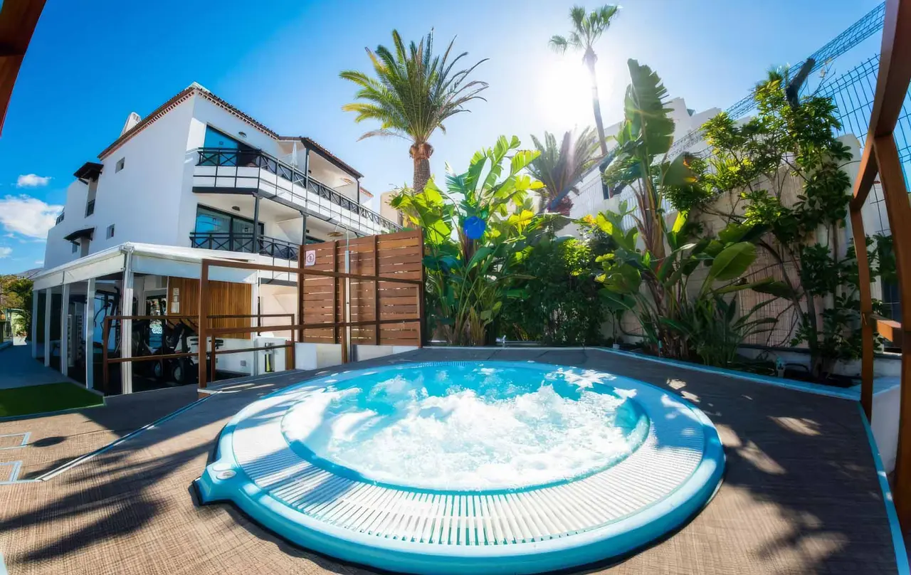Jacuzzi circular con agua burbujeante en una terraza exterior rodeada de vegetación tropical, palmeras y edificios