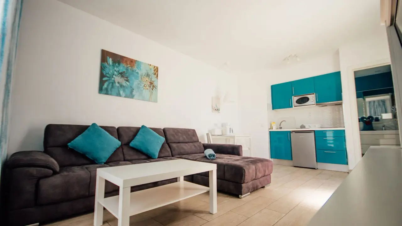 Interior de una sala de estar con sofá marrón en forma de L, cojines turquesa, mesa de centro blanca, cocina americana