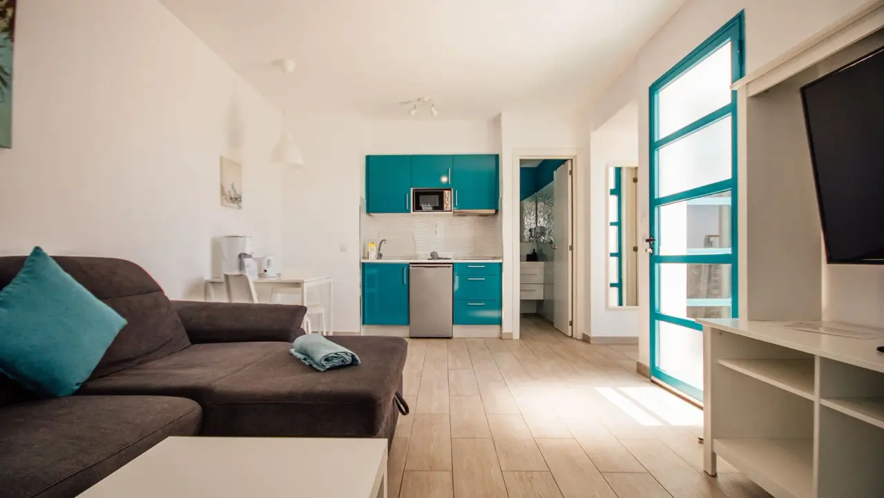 Interior de un bungalow con sala de estar, sofá marrón, cocina americana con muebles turquesa al fondo y puerta
