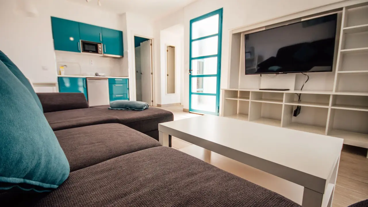 Interior de un bungalow con sofá marrón en primer plano, mesa blanca, estantería con televisor y cocina americana