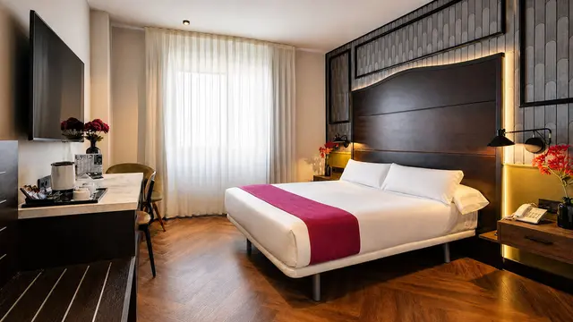Vista de una junior suite con cama doble grande vestida de blanco con una banda central magenta, cabecero de madera