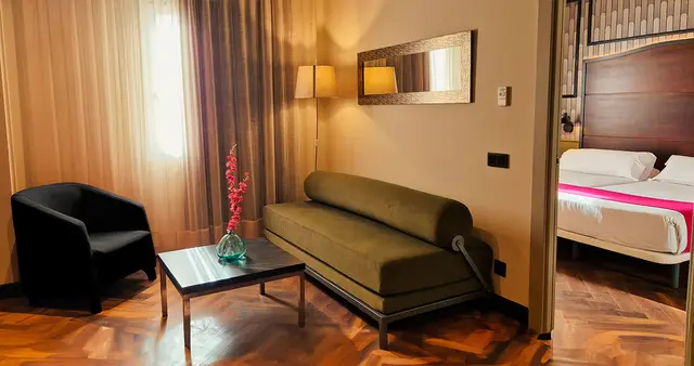 Vista de una sala de estar en una suite de hotel con un sofá verde, un sillón negro, una mesa de centro con un jarrón