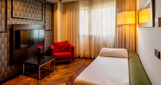 Vista de una junior suite con cama individual vestida de blanco, butaca roja, mesa de centro con jarrón de flores