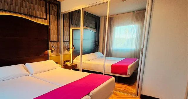Vista de una junior suite con dos camas individuales, cabeceros de madera oscura con iluminación indirecta, mesitas
