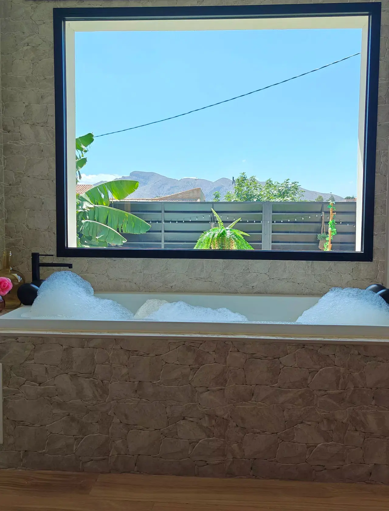 Jacuzzi interior con agua espumosa, situado frente a una gran ventana que muestra un jardín con plantas, una valla