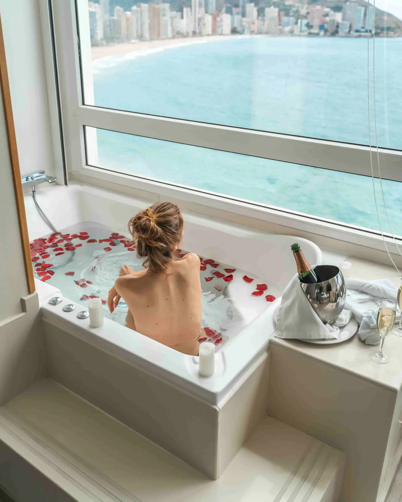 Mujer de espaldas dentro de una bañera blanca con agua y pétalos de rosa rojos, en un spa privado con vista panorámica