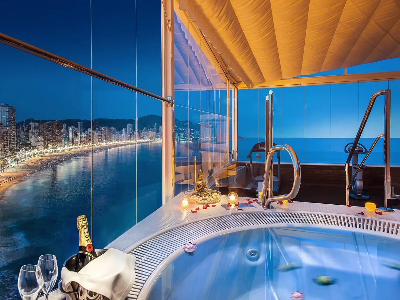 Vista nocturna de un spa privado con jacuzzi iluminado en primer plano, rodeado de pétalos de rosa, velas