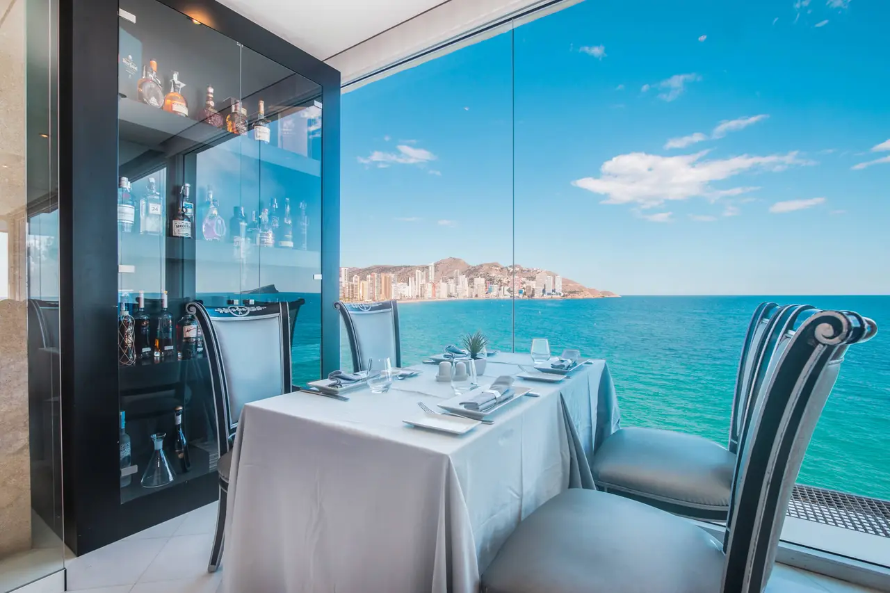 Vista desde el interior de un comedor con mesa puesta para dos personas, sillas elegantes y un aparador con botellas