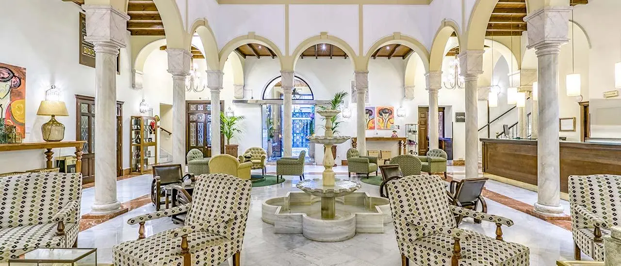 Vista de un amplio vestíbulo o sala común con arcos de piedra, columnas, suelo de mármol, sillones tapizados en blanco