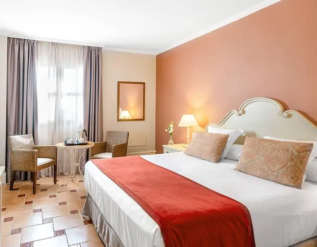 Vista de una suite de hotel con una cama grande con cabecero blanco y colcha roja, dos mesitas de noche con lámparas