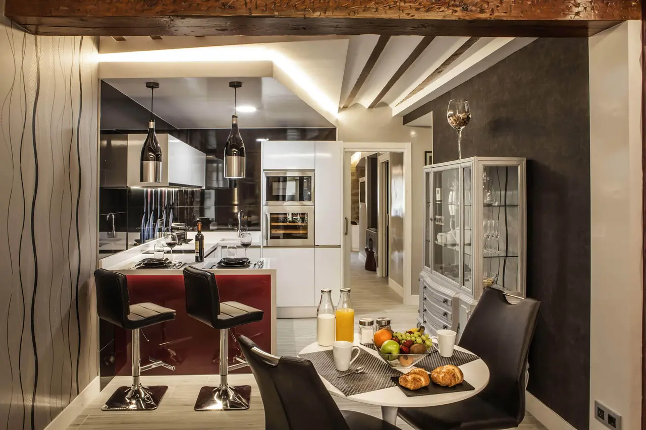 Vista de una suite con zona de cocina moderna, isla con taburetes negros, mesa redonda con desayuno y sillas de cuero