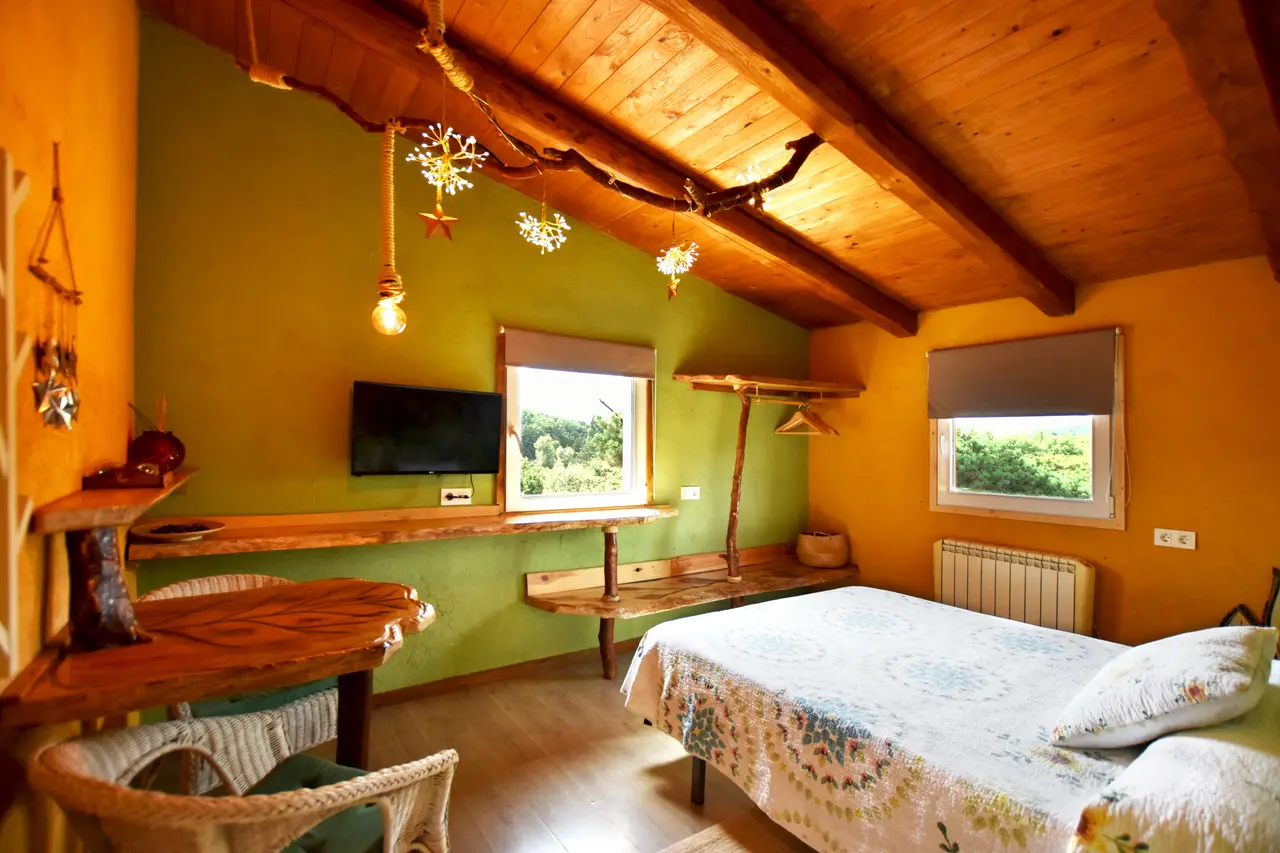 Habitación acogedora con cama, escritorio de madera, sillas de mimbre y techo abuhardillado de vigas vistas.