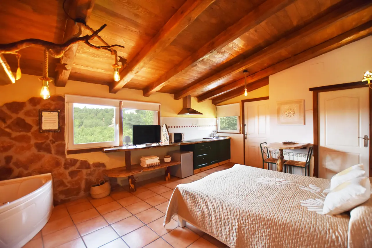 Habitación rústica con techo de madera, cama grande, bañera, cocina y vistas al bosque a través de las ventanas.