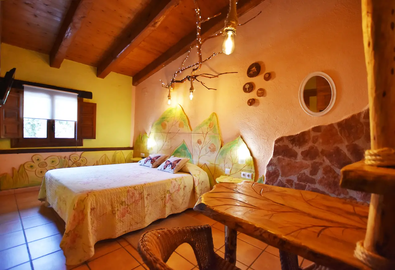 Habitación rústica con cama doble, cabecero de hojas pintadas, mesa de madera y paredes de piedra y coloridas.