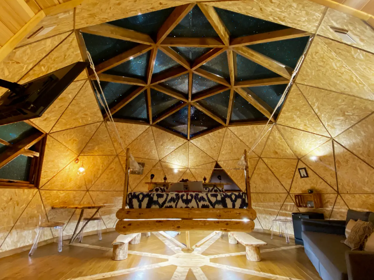 Interior de un iglú geodésico de madera con techo estrellado, cama redonda central y mobiliario rústico.