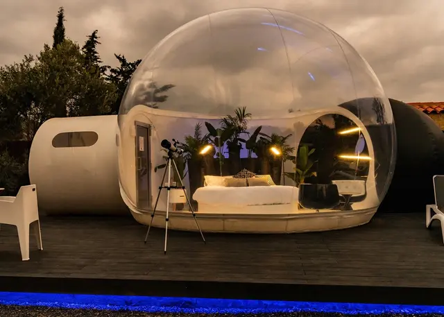 Burbuja transparente iluminada con cama interior, plantas y telescopio en terraza exterior al atardecer.