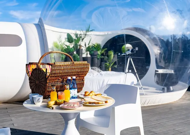 Mesa con desayuno y cesta de picnic en terraza exterior frente a una burbuja transparente de alojamiento.