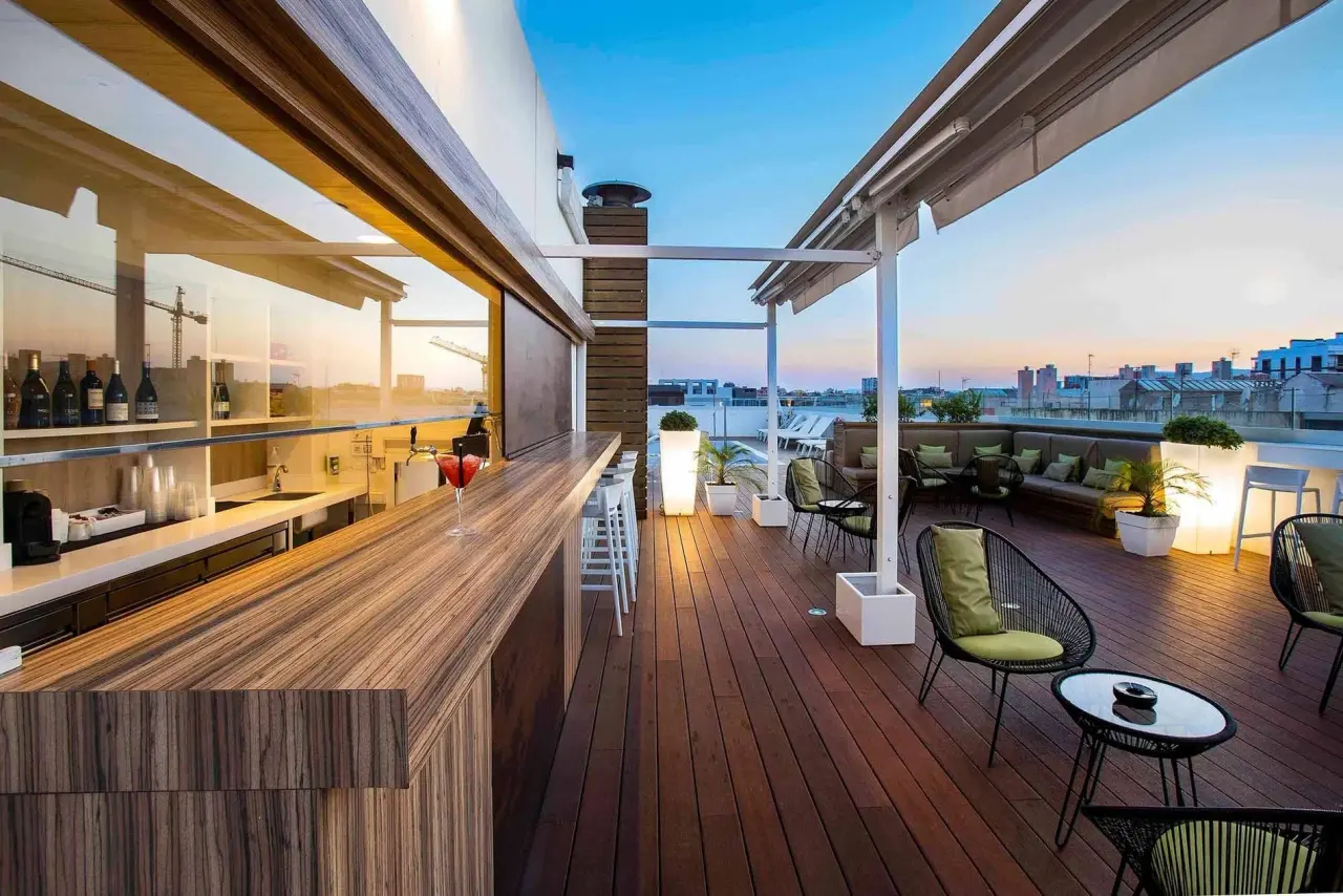 Terraza exterior de un hotel al atardecer con suelo de madera, una barra de bar con estantes de botellas y copas